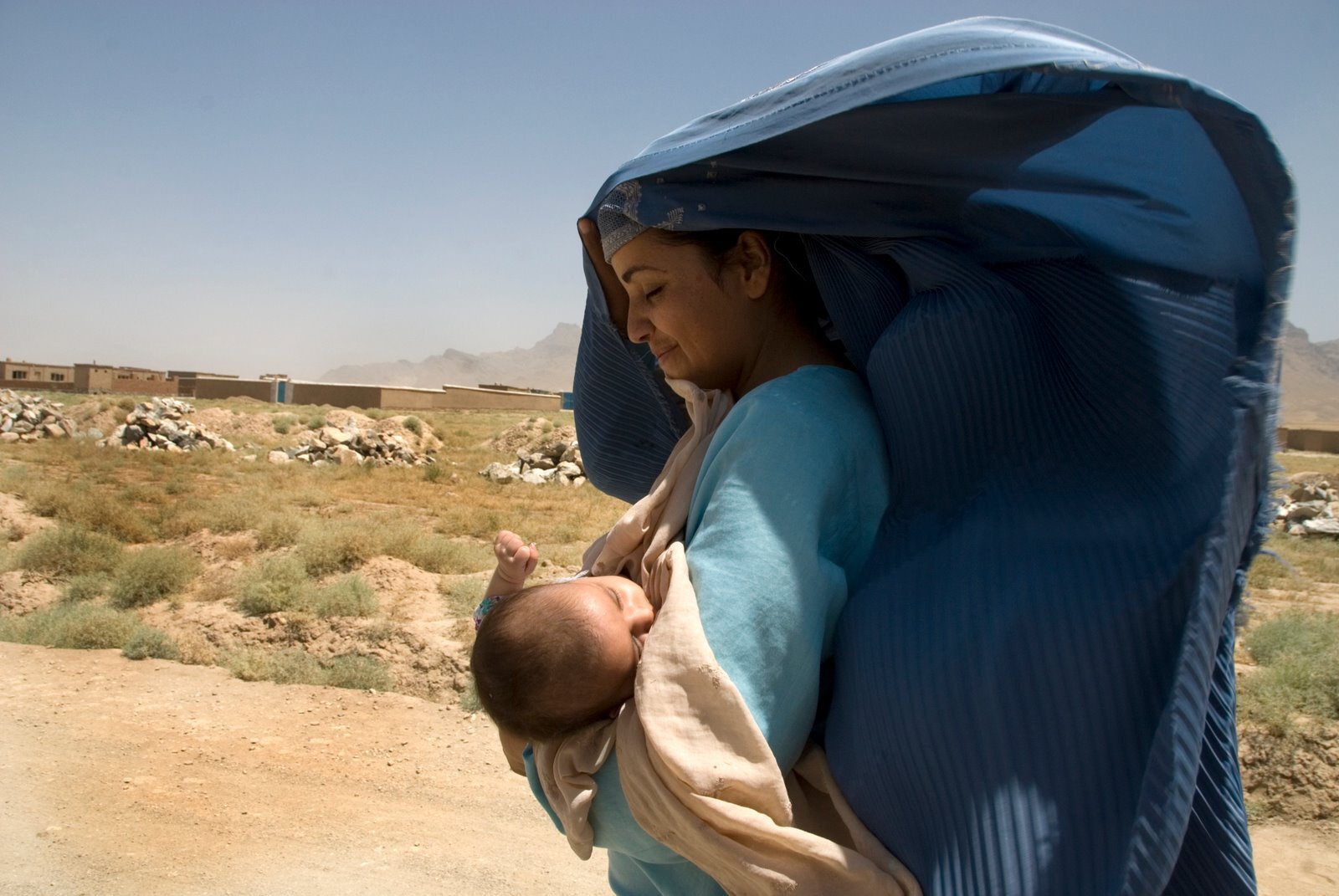 En kvinne holder sitt barn i armene. Hun har på seg et blått sjal som flagrer i vinden